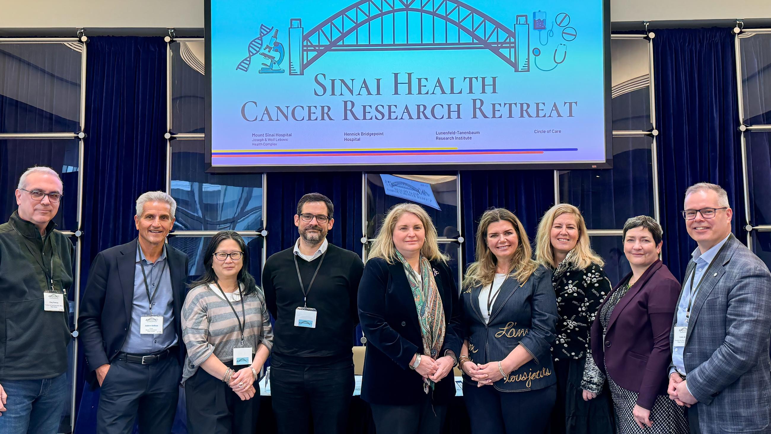A group of nine health-care professionals standing together in a single row, facing the camera smiling. They are wearing business attire. They are standing under a screen with an image of a bridge and the words, Sinai Health Cancer Research Retreat