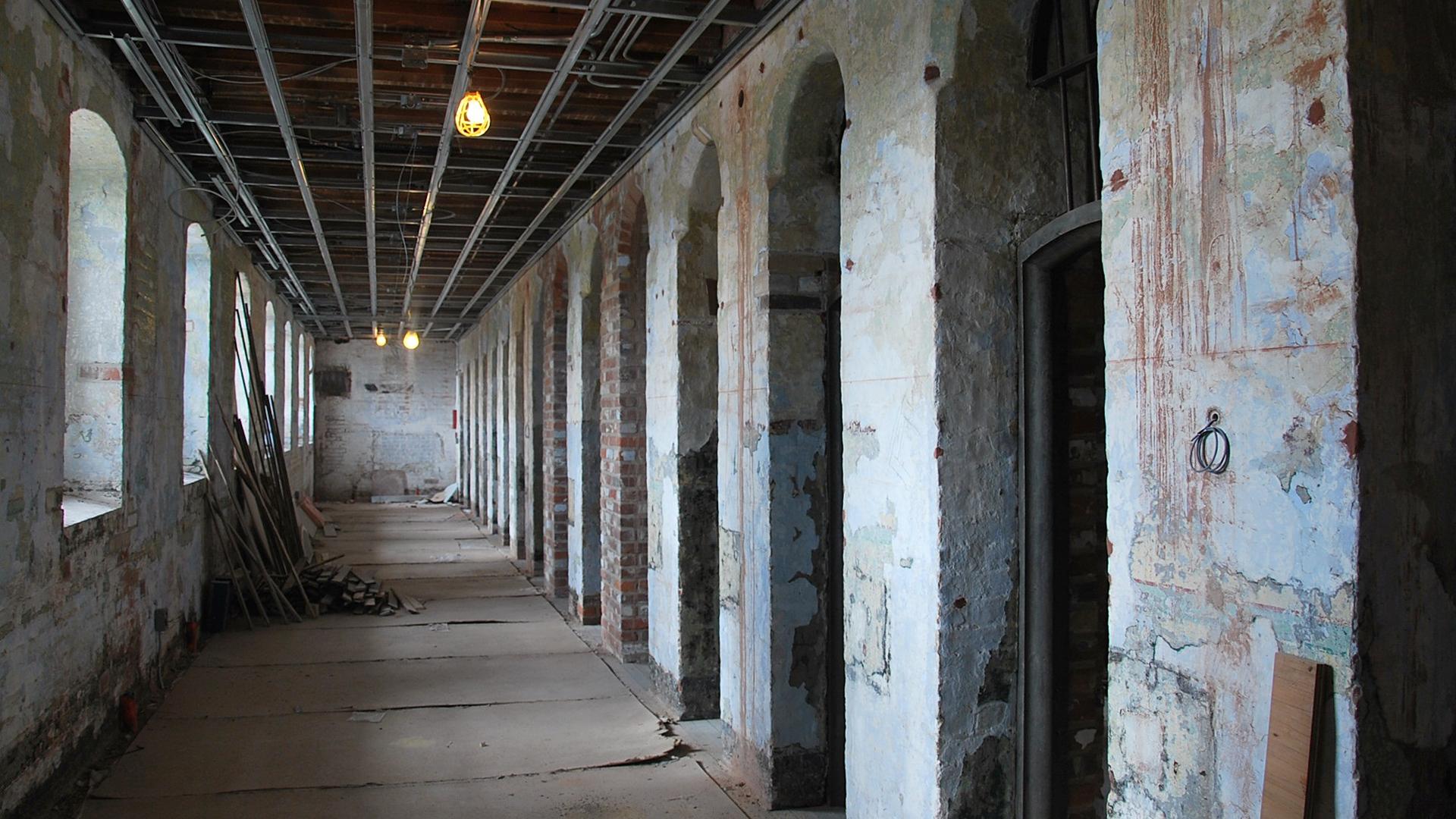 A view of the Don Jail cell block during renovation, looking down the length of a corridor. There are arched door frames on the right side and windows on the left side. The hall is under construction and unfinished.
