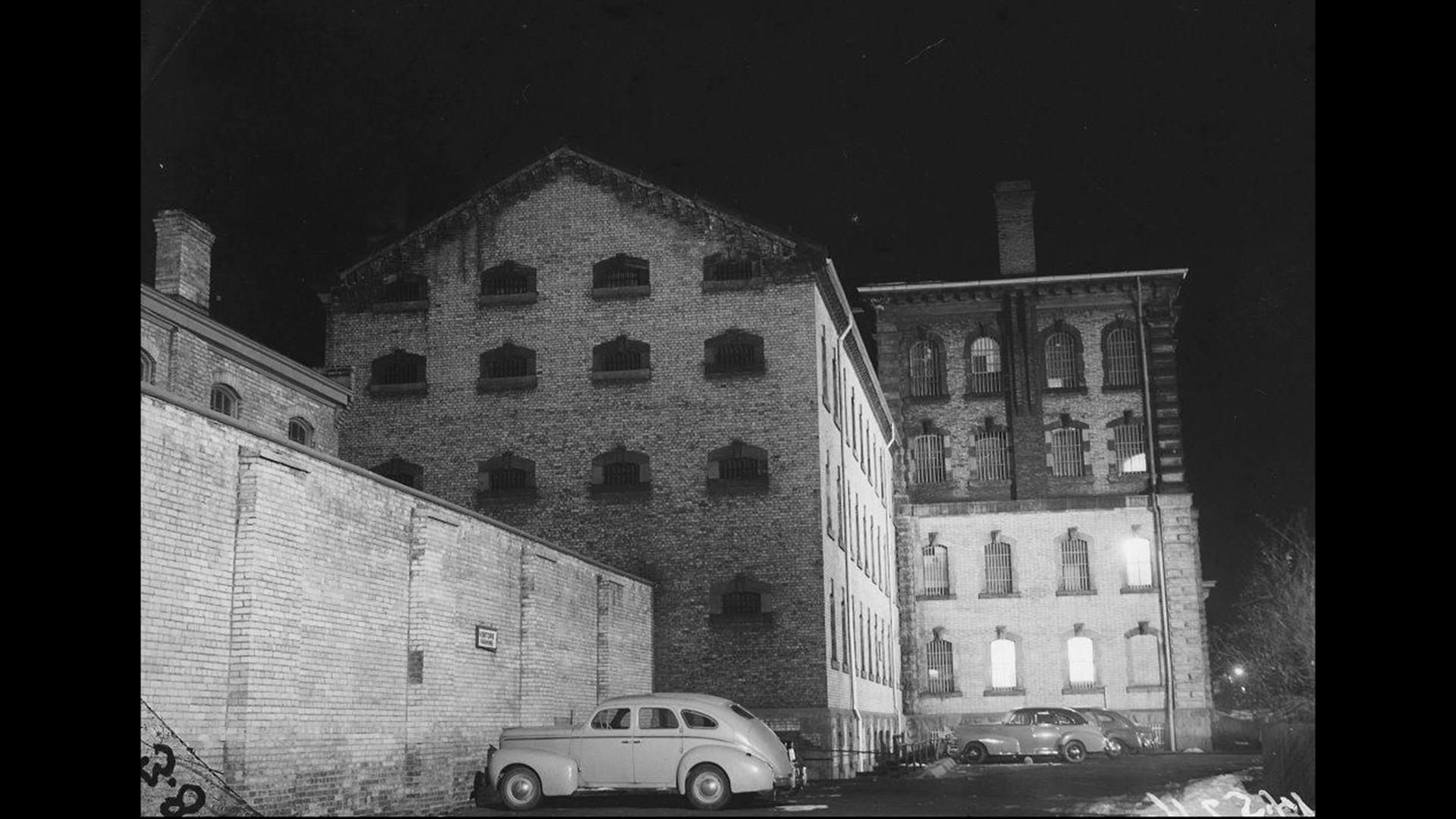 Night image of the Don Jail around 1952. The building is lit up by floodlights. It is four storeys and made of light brick. The bars on the windows are visible. There are a few cars in front of the building. The photo is in black and white.