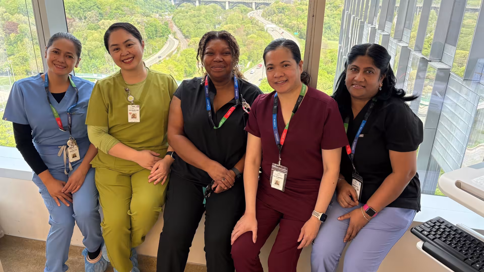 A group of four hospital employees wearing scrubs. They are sitting in a row in front of a window with a view of a green leafy valley behind them. They are looking at the camera and smiling