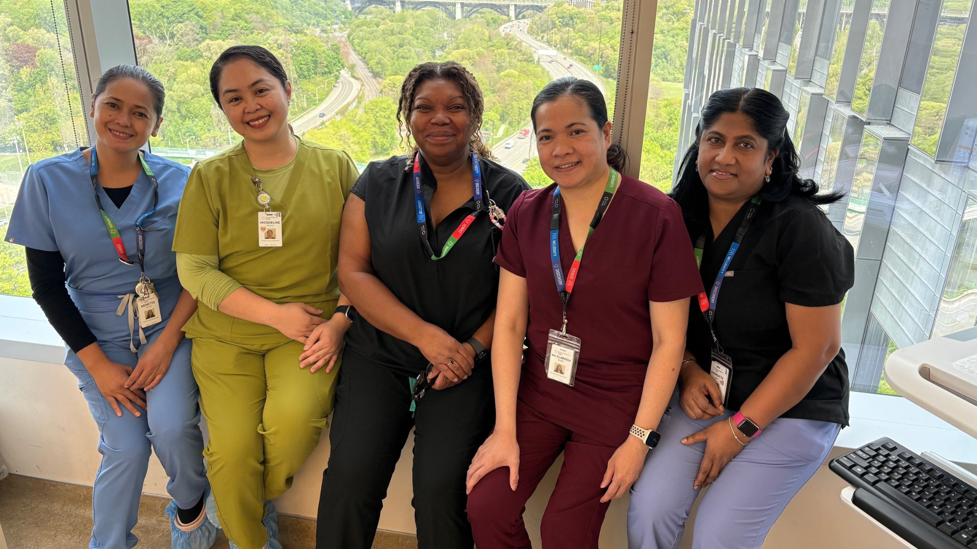 A group of four hospital employees wearing scrubs. They are sitting in a row in front of a window with a view of a green leafy valley behind them. They are looking at the camera and smiling