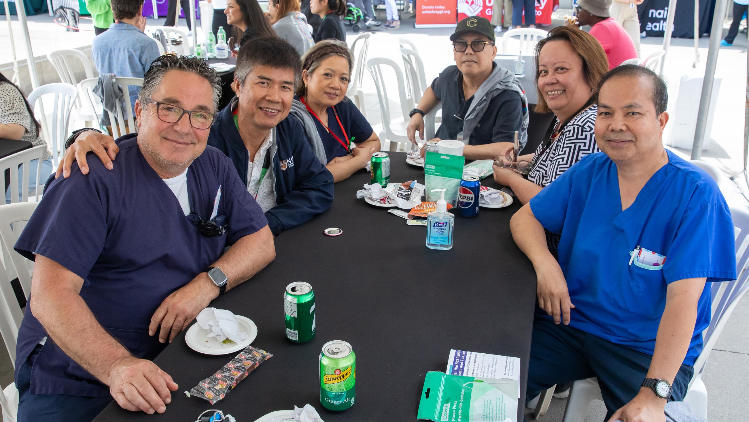 A group of six colleagues smiling and sitting together around a table at the barbeque. 