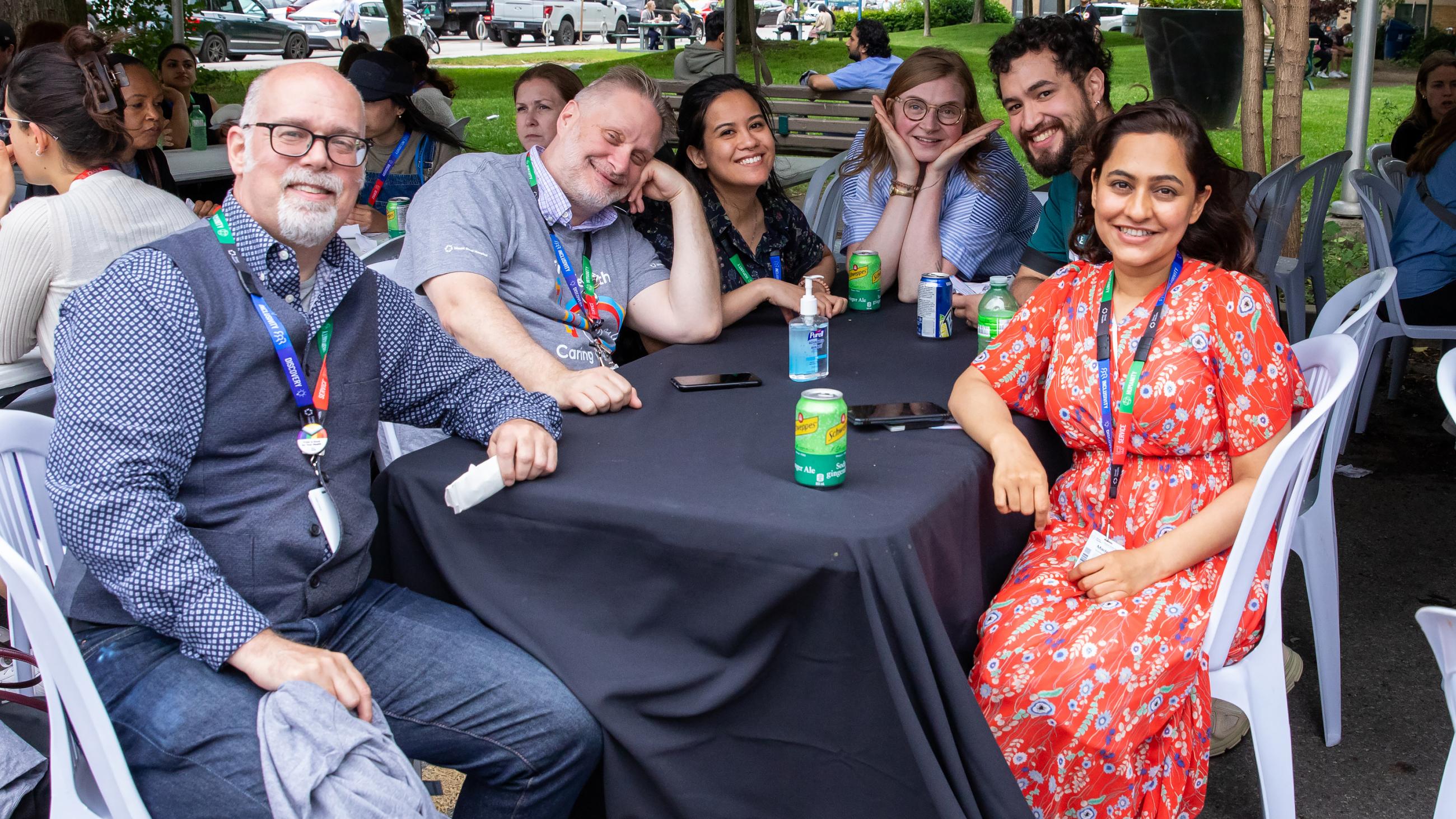 A group of six colleagues smiling and sitting together around a table at the barbeque.