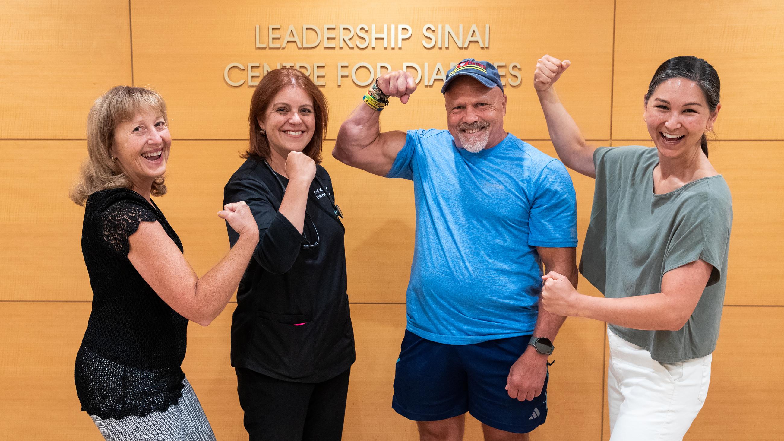 Four people pose in front of a wall that reads "Leadership Sinai Centre for Diabetes". A muscular man in a blue shirt and shorts flexes his arm, beside three smiling women - one in scrubs, one in a black lace top, and one in a light green top - who are also playfully flexing their arms.