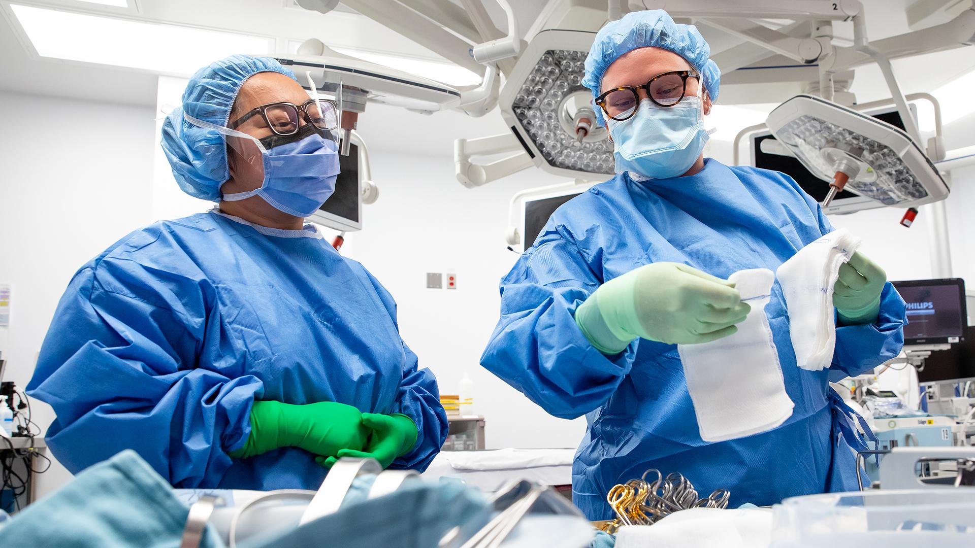 Two nurses stand in front of a table of surgical instruments in an Operating room. The nurses are wearing scrubs, including blue gown, gloves, caps and face shields. One nurse is holding out surgical sponges while the other watches