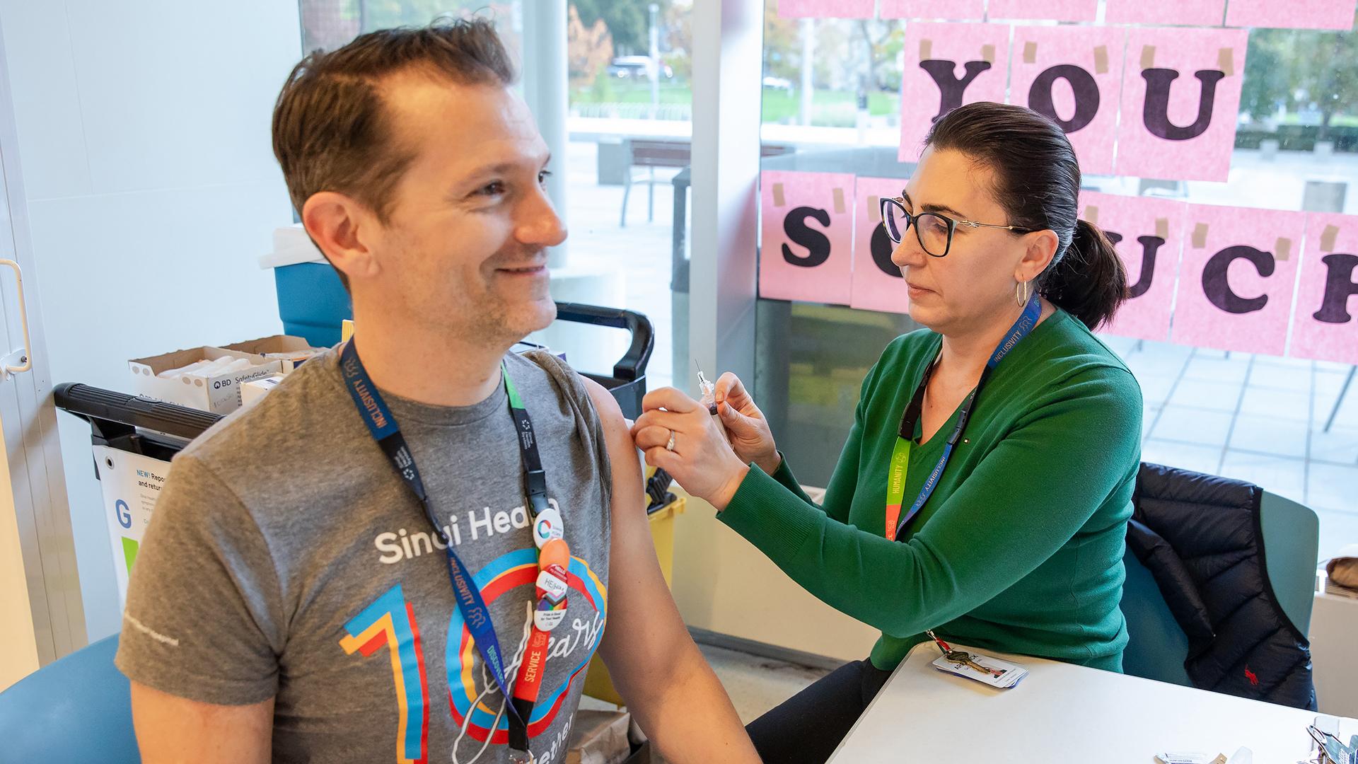A woman prepares to give a vaccination to a man. They are both sitting at a table that is in front of a window.