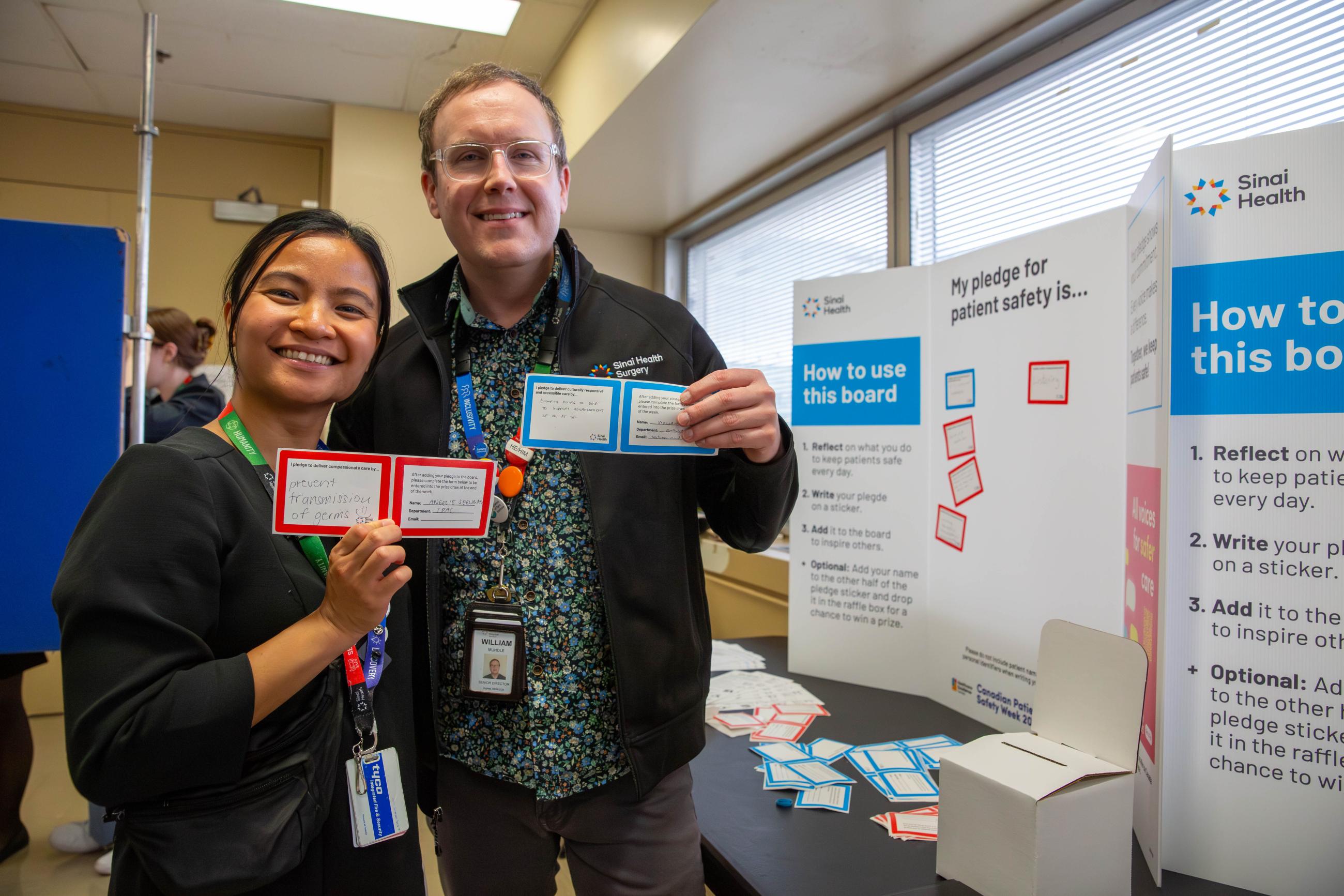 Two people standing next to a table with a display board on it. They are holding up stickers that they have written on that will be stuck on the display board.