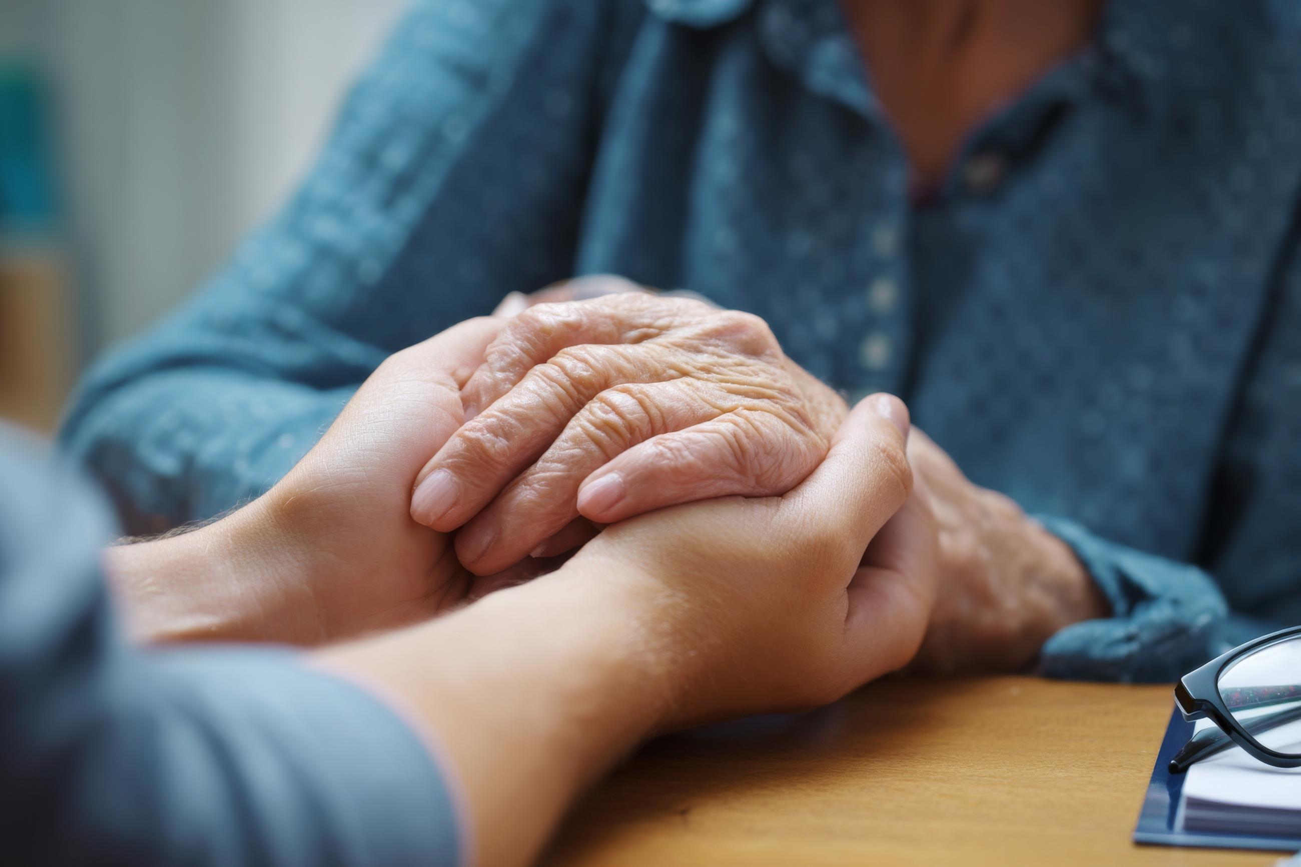 A person gently holding the hands of an older adult across a table, conveying comfort and support.
