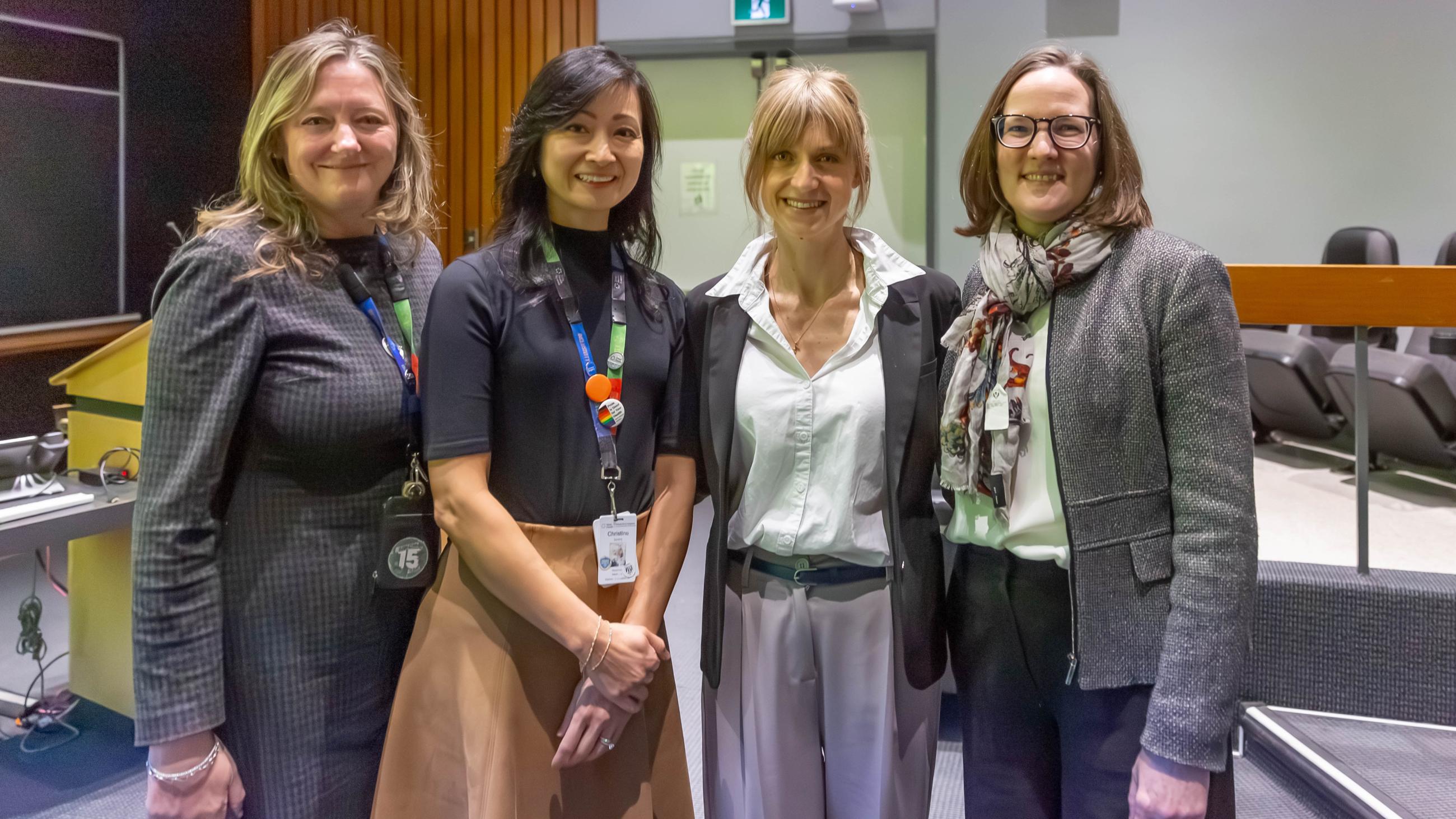 A group of four women standing in a line, side-by-side. They are looking at the camera, smiling. They are in an auditorium