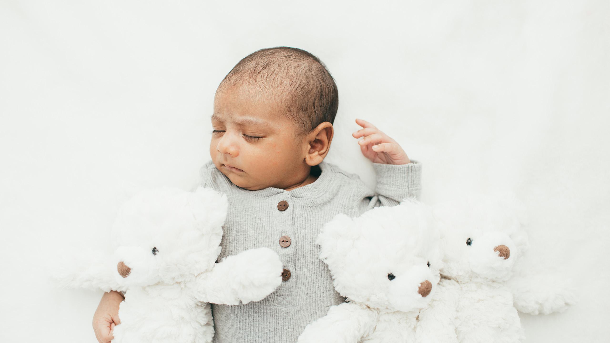 Infant boy wearing a light grey shirt sleeping with white teddy bears around him
