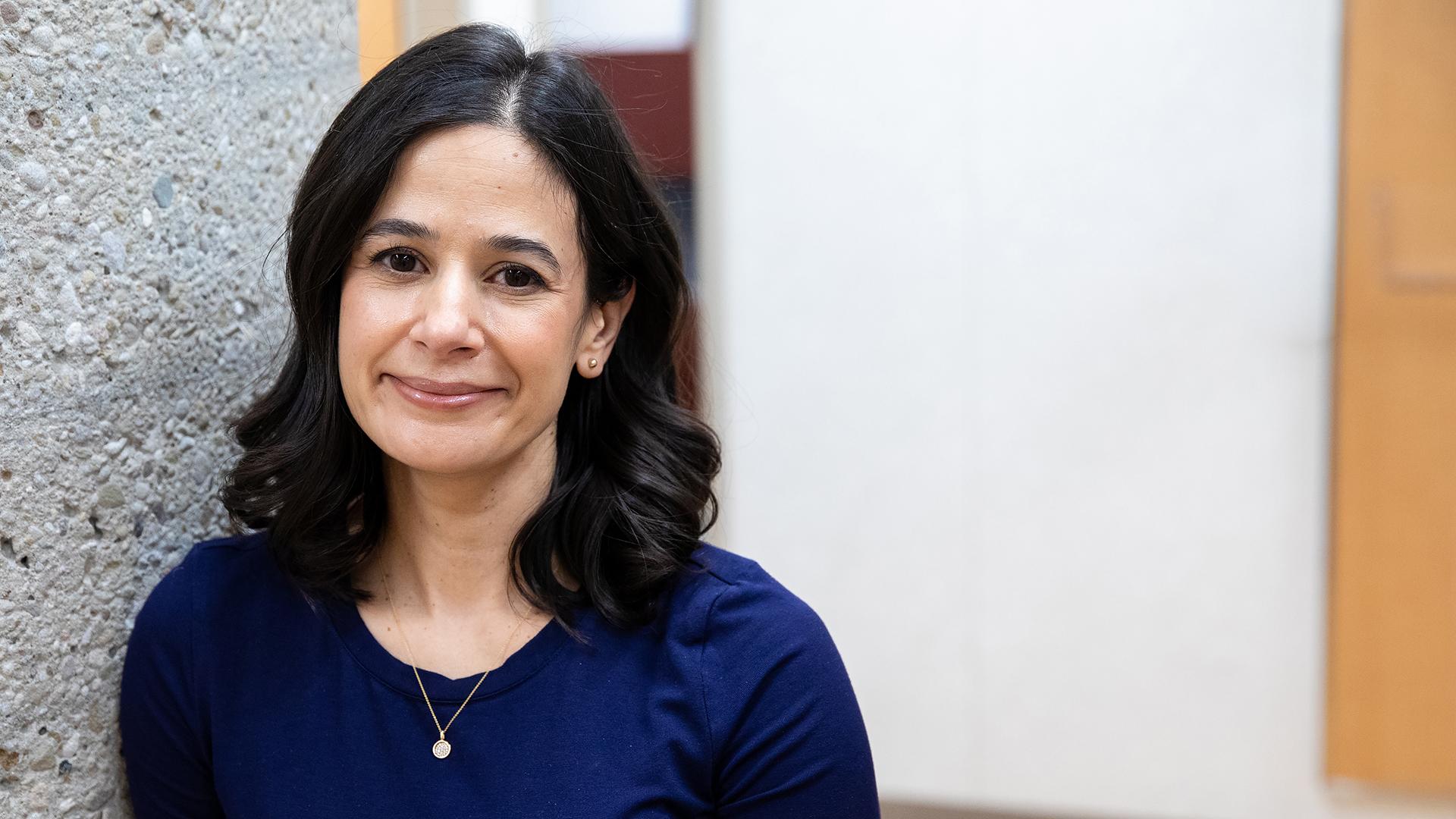 Dr. Amanda Selk portrait. She is standing next to a textured concrete pillar and looking at the camera, smiling.
