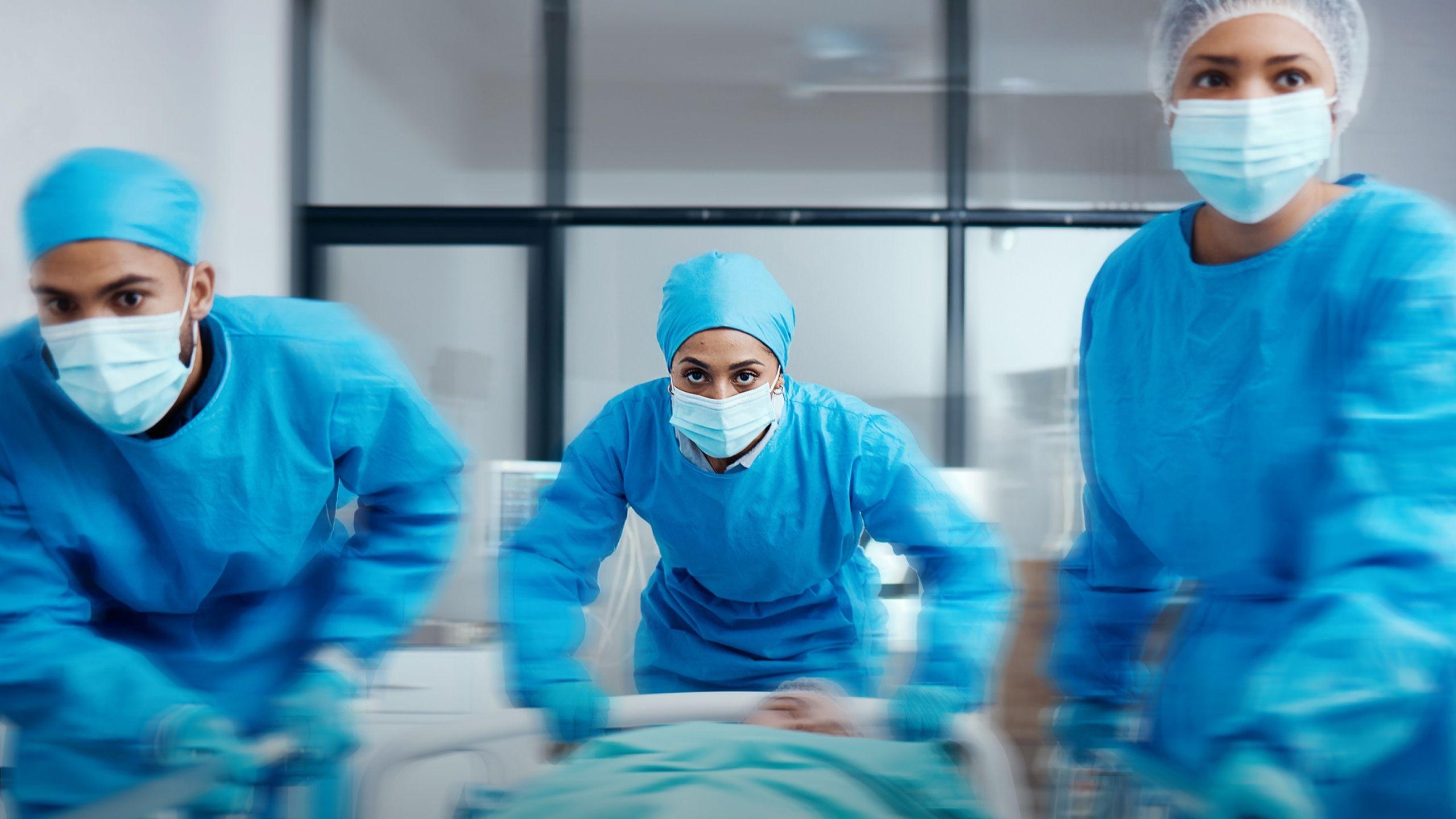 Three clinicians in blue surgical attire rush a patient down a hospital corridor, conveying urgency and coordinated emergency care.