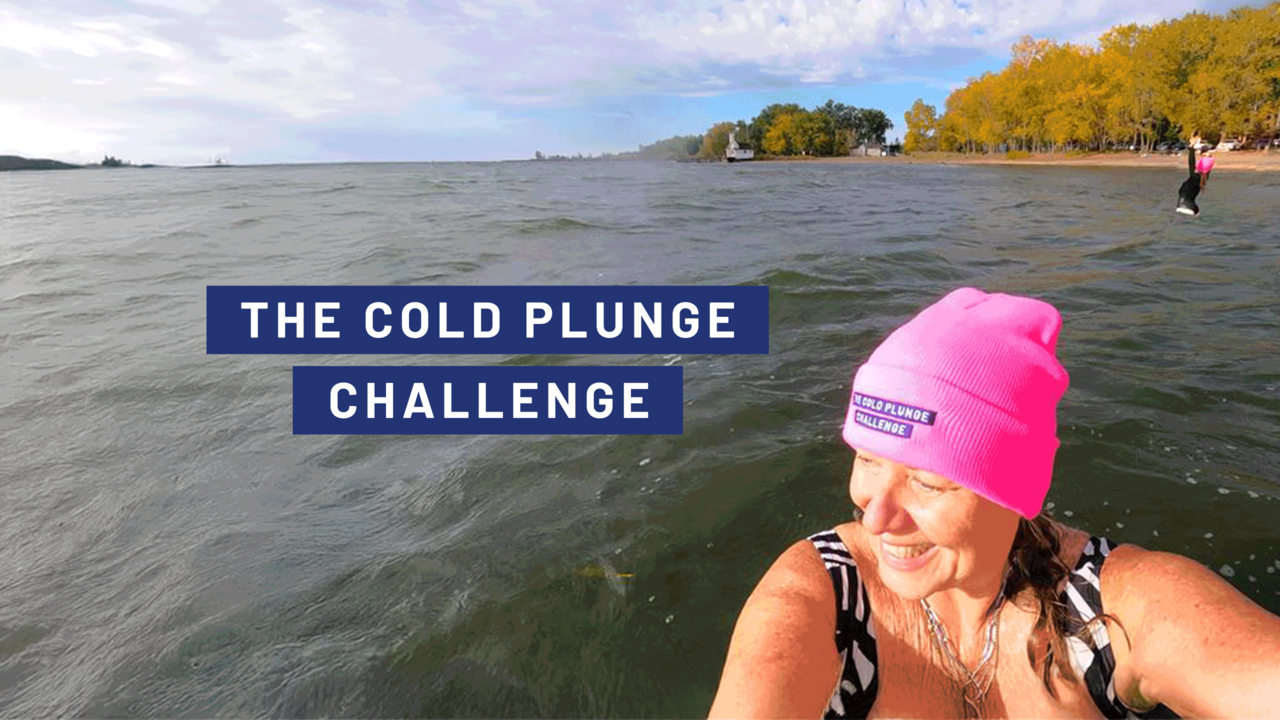 A person wearing a bright pink winter hat stands in cold lake water near a shoreline lined with autumn trees. Another participant is visible farther out on the right. Text over the water reads “The Cold Plunge Challenge” against a blue background.