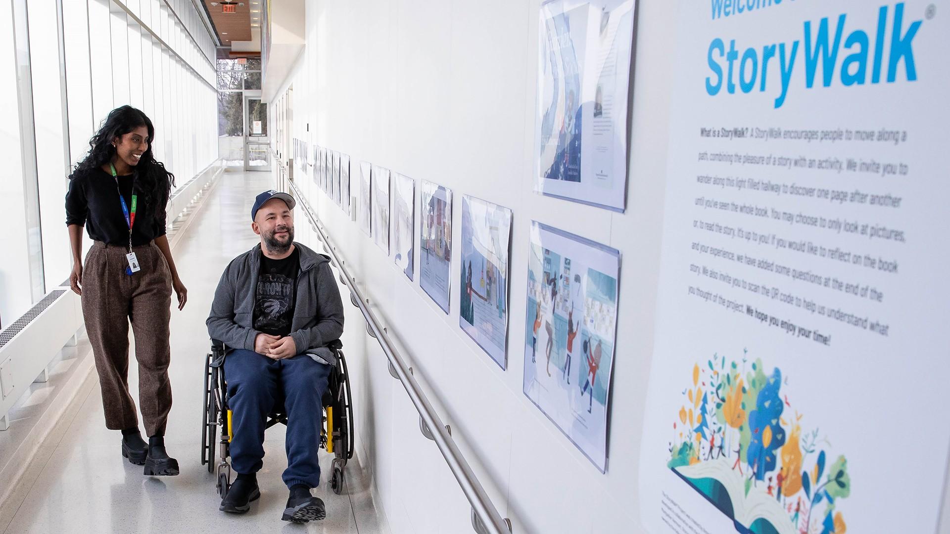 A patient using a wheelchair moves along a hospital hallway with an occupational therapist, viewing the Story Walk panels displaying illustrated book pages mounted on the wall at Hennick Bridgepoint Hospital.