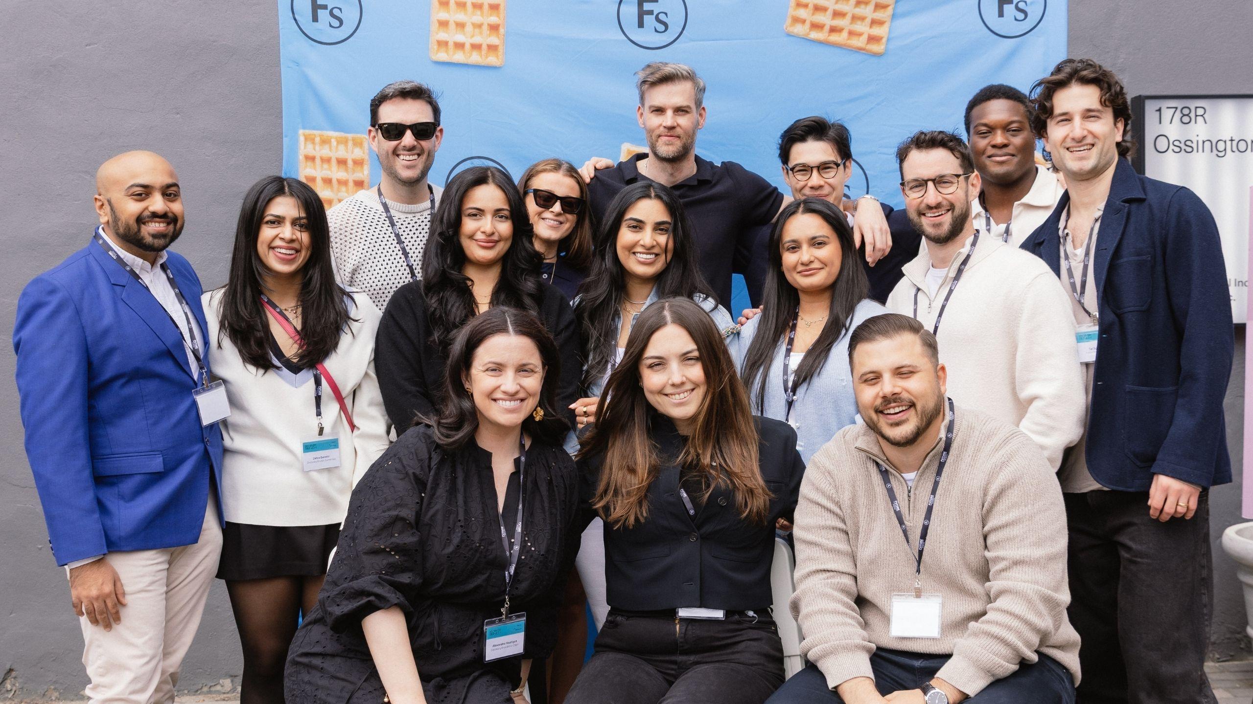 Fifteen guests arranged in three rows in front of the Discovery Brunch event backdrop, smiling with their arms around each other. 