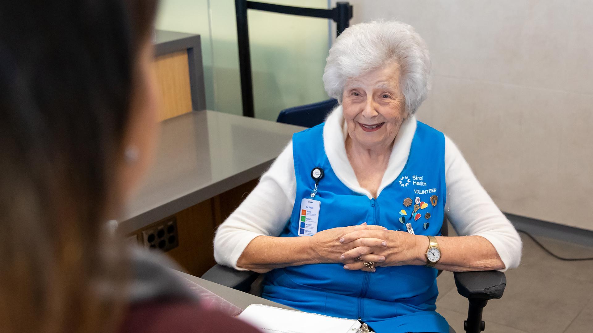 A woman sits at an information desk at a hospital greeting a patient with a smile on her face. 