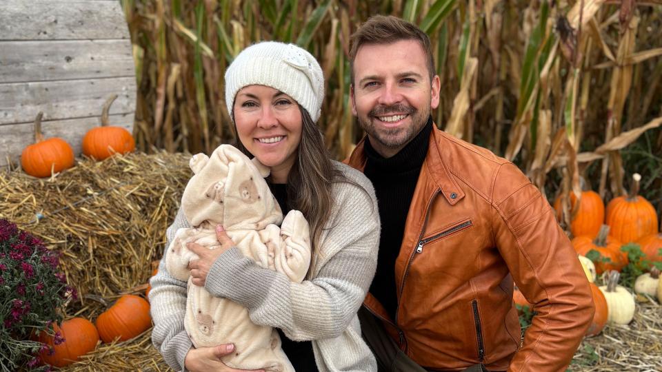 woman holding baby sits next to a man in against a fall backdrop