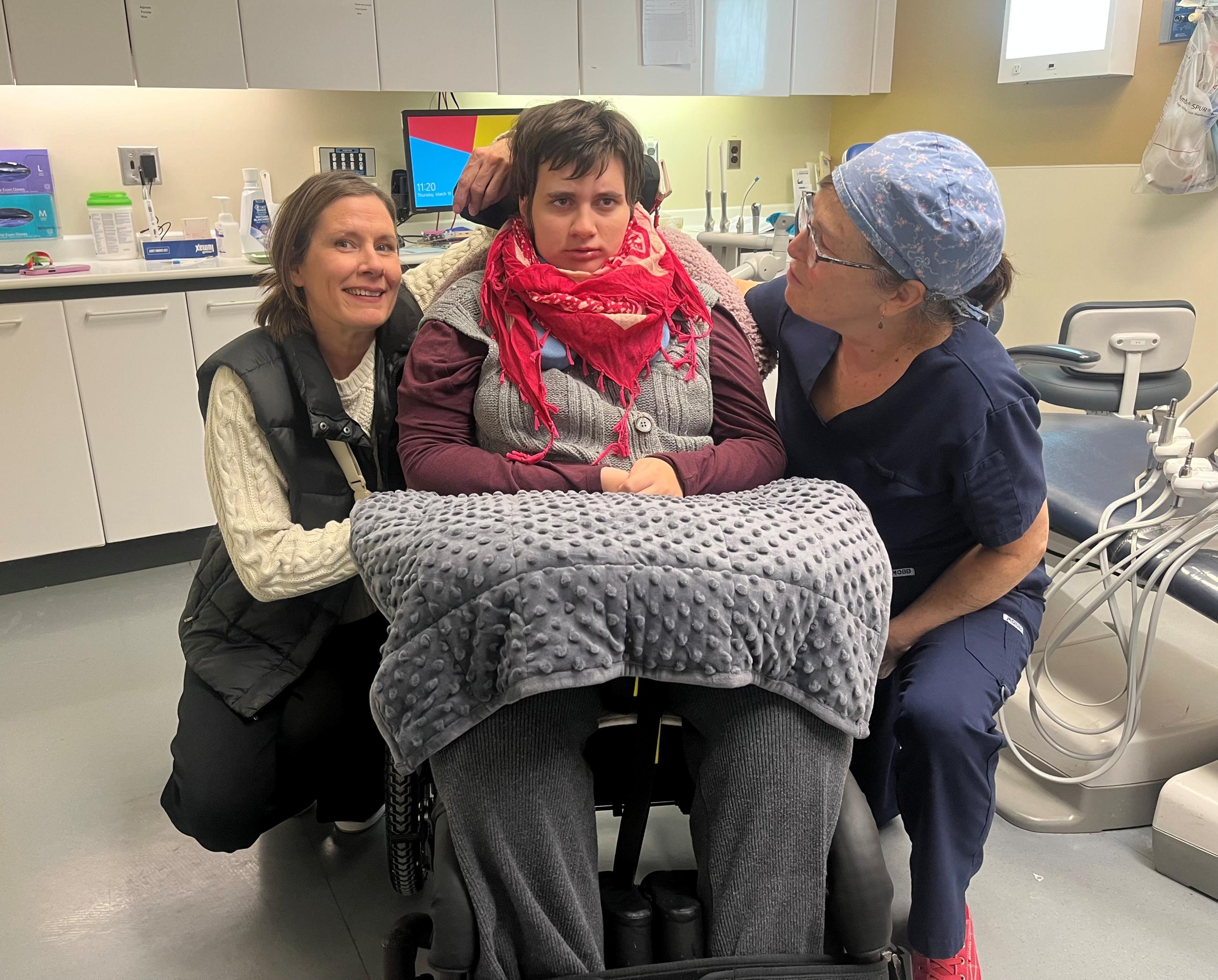Young woman sits in a wheelchair in a dental clinic, with her dentist and mother smiling beside her