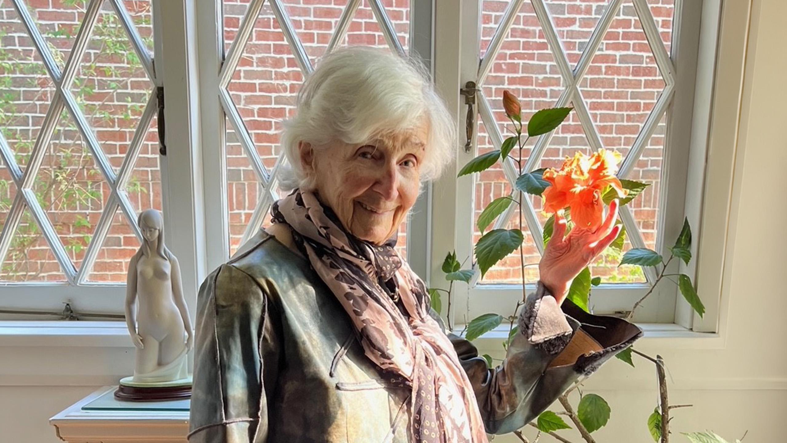 elderly woman stands by a window holding a flower