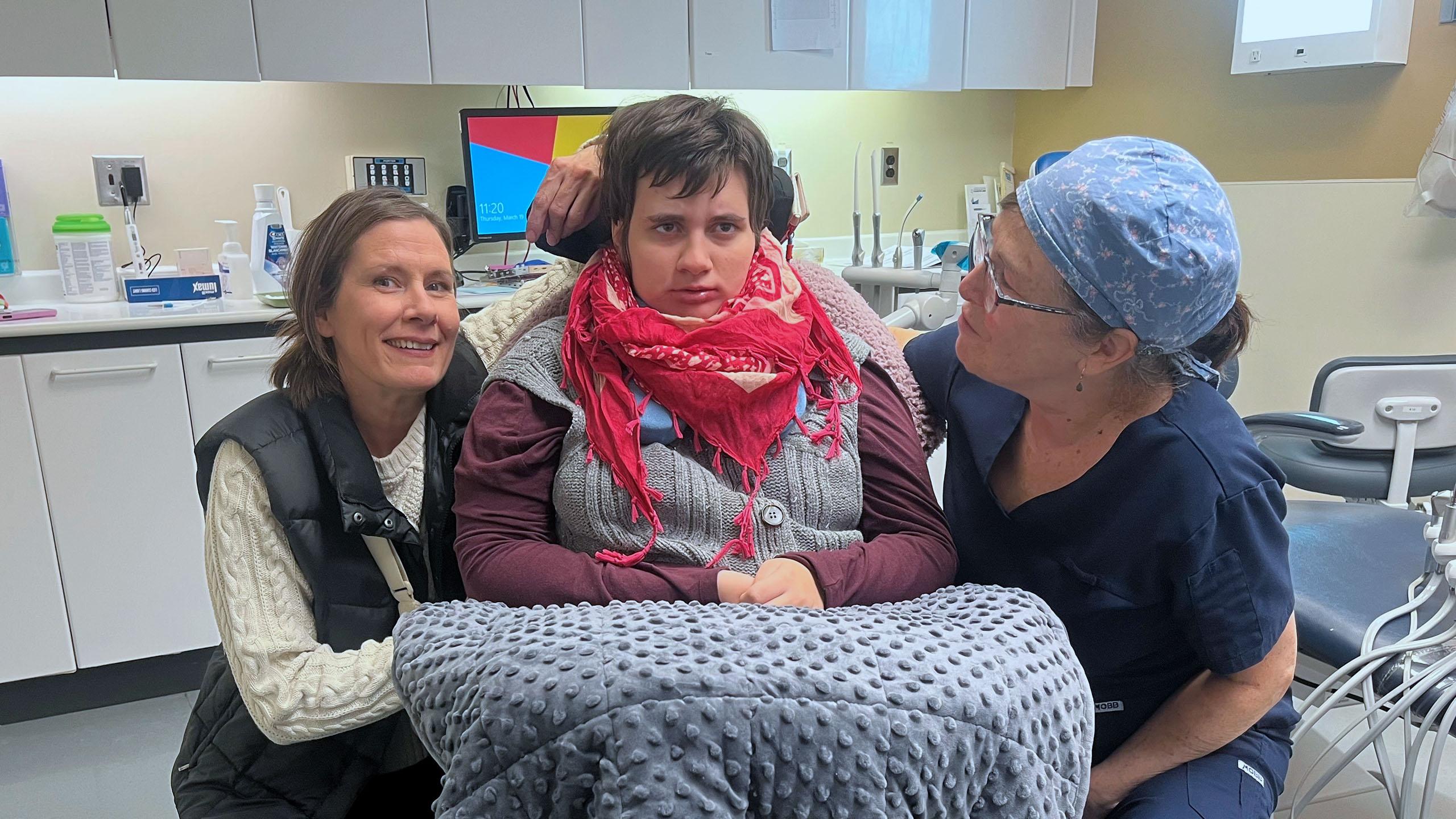Young woman sits in a wheelchair in a dental clinic, with her dentist and mother smiling beside her