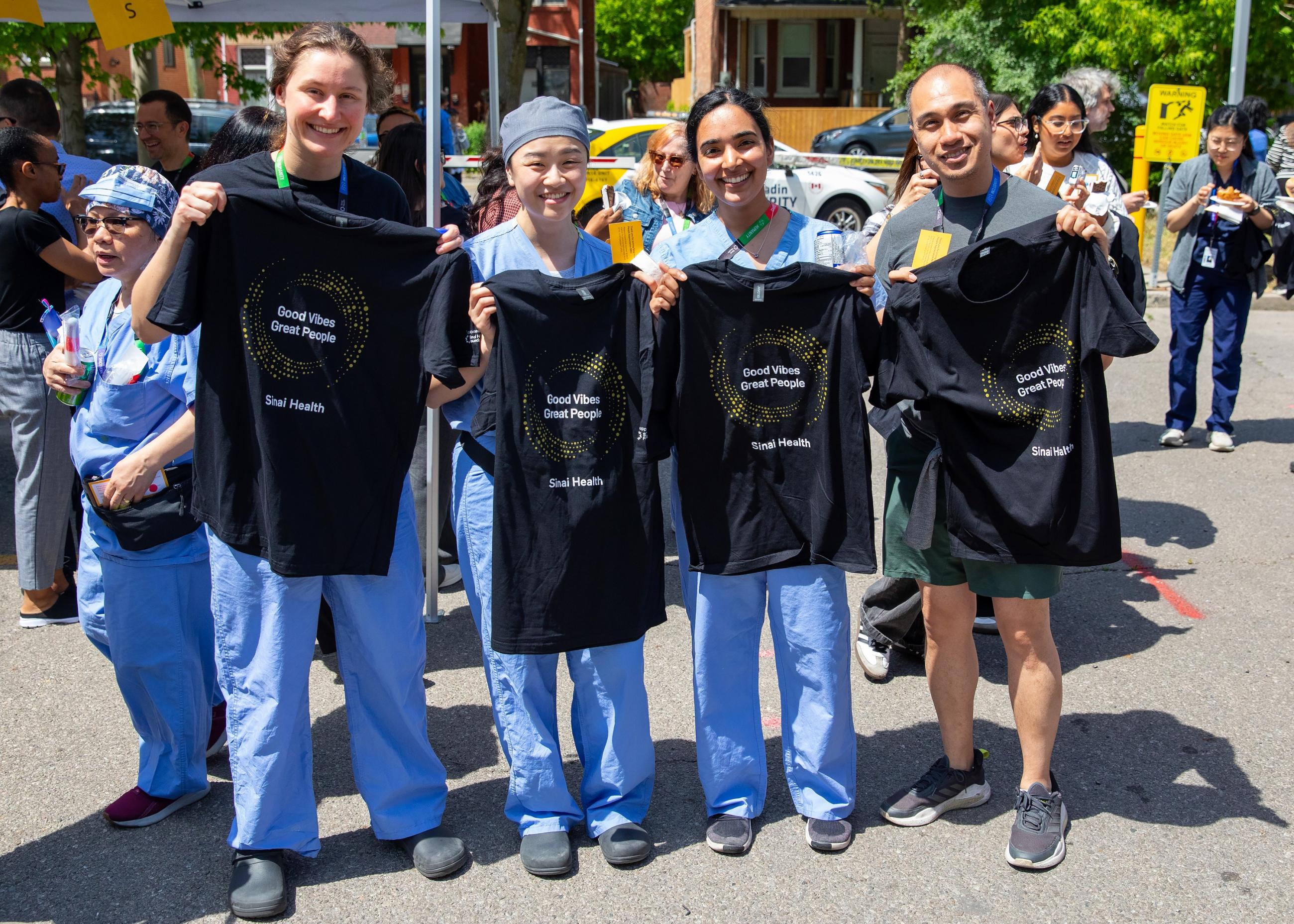 Four Sinai Health employees stand outdoors at the People Appreciation BBQ, smiling and holding black “Good Vibes Great People” T-shirts.
