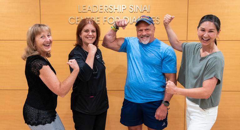 Four people pose in front of a wall that reads "Leadership Sinai Centre for Diabetes". A muscular man in a blue shirt and shorts flexes his arm, beside three smiling women - one in scrubs, one in a black lace top, and one in a light green top - who are also playfully flexing their arms.