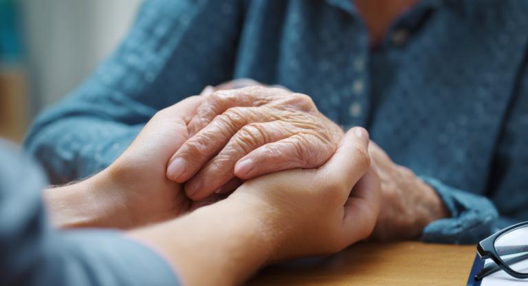 A person gently holding the hands of an older adult across a table, conveying comfort and support.