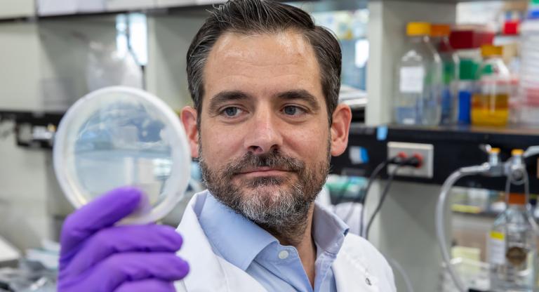 scientist examining a petri dish, he is wearing a lab coat and purple gloves