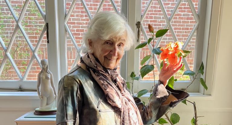 elderly woman stands by a window holding a flower