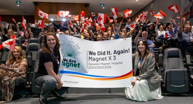 Two people holding a banner celebrating the Magnet recognition in an auditorium with people cheering behind them waving Canadian flags.