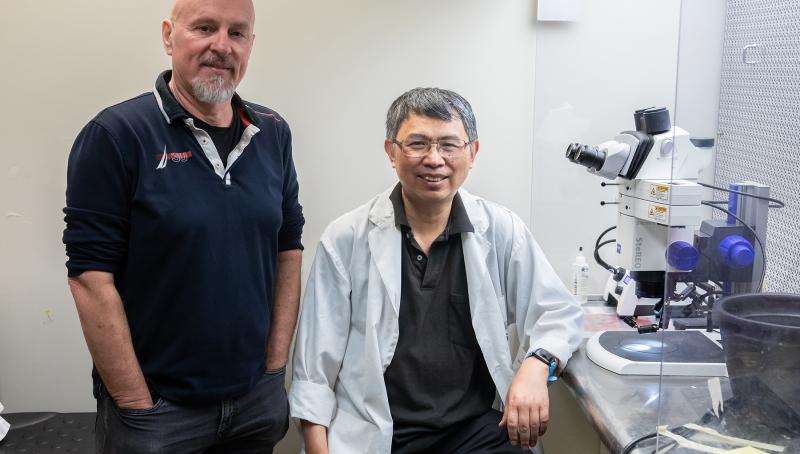 Two researchers pictured in the laboratory next to a microscope. One researcher. A Caucasian man is standing with hands in pockets and the other, a bespectacled Asian man, is seated and wears a lab coat.