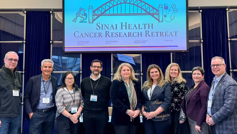 A group of nine health-care professionals standing together in a single row, facing the camera smiling. They are wearing business attire. They are standing under a screen with an image of a bridge and the words, Sinai Health Cancer Research Retreat