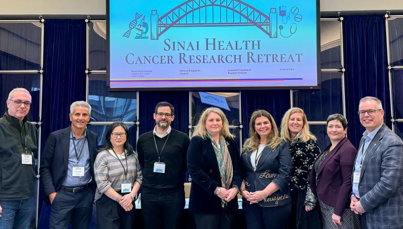 A group of nine health-care professionals standing together in a single row, facing the camera smiling. They are wearing business attire. They are standing under a screen with an image of a bridge and the words, Sinai Health Cancer Research Retreat