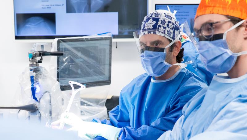 Dr. Jesse Wolfstadt in a blue gown and mask using advanced robotic equipment in a sterile operating room.