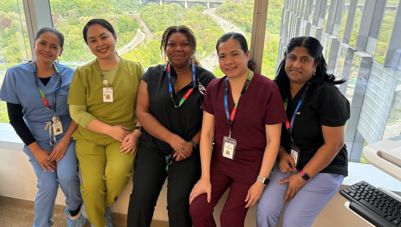 A group of four hospital employees wearing scrubs. They are sitting in a row in front of a window with a view of a green leafy valley behind them. They are looking at the camera and smiling