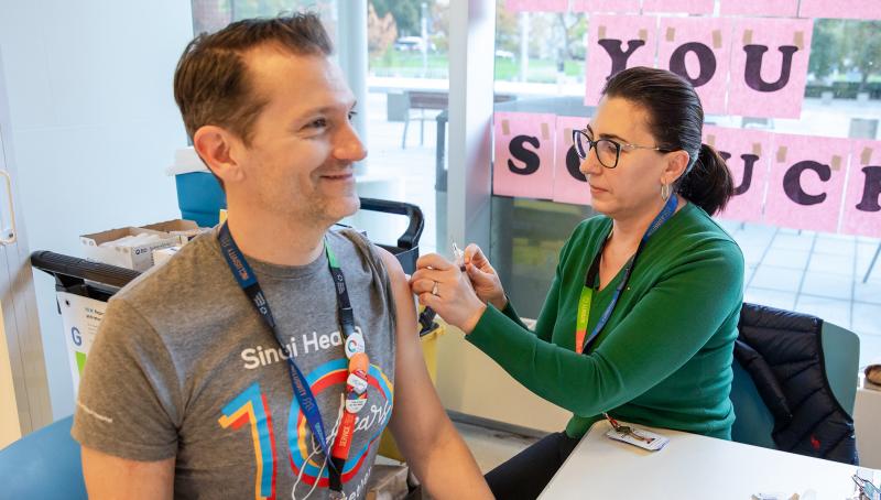 A woman prepares to give a vaccination to a man. They are both sitting at a table that is in front of a window.
