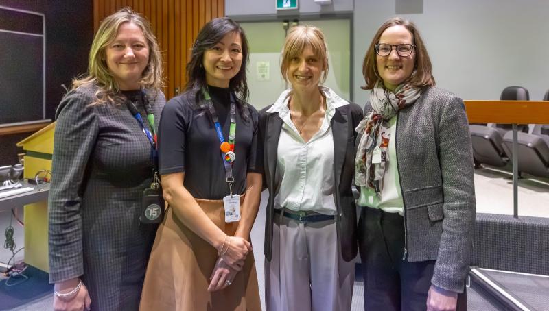 A group of four women standing in a line, side-by-side. They are looking at the camera, smiling. They are in an auditorium