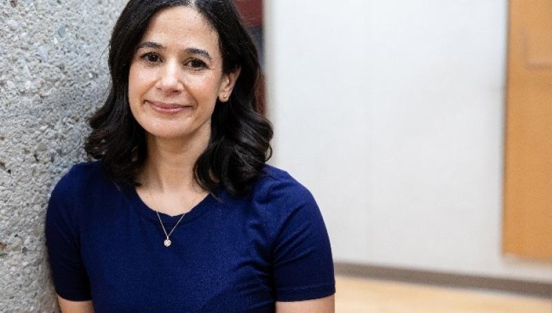 Dr. Amanda Selk stands indoors beside a textured concrete pillar, smiling softly at the camera while wearing a navy blue top and small gold necklace. The background shows a bright hallway with light flooring and neutral walls.