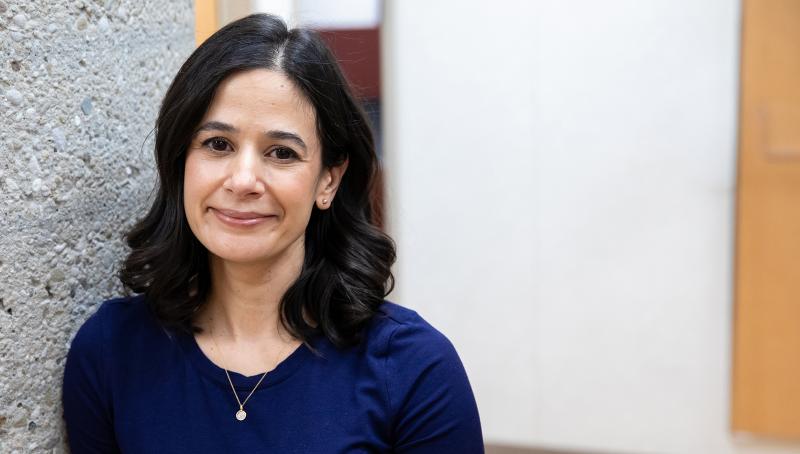 Dr. Amanda Selk portrait. She is standing next to a textured concrete pillar and looking at the camera, smiling.