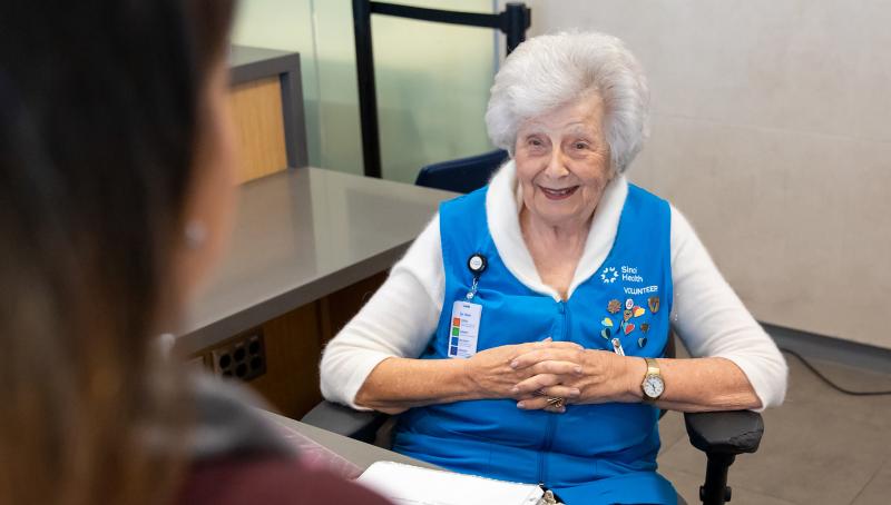 A woman sits at an information desk at a hospital greeting a patient with a smile on her face. 