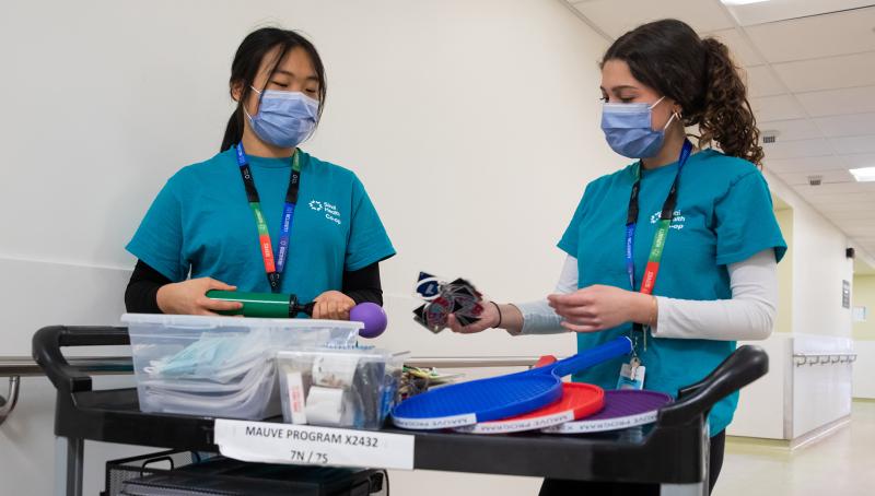 Two Sinai Health co-op students wearing teal scrubs and masks stand beside a cart stocked with recreational items.