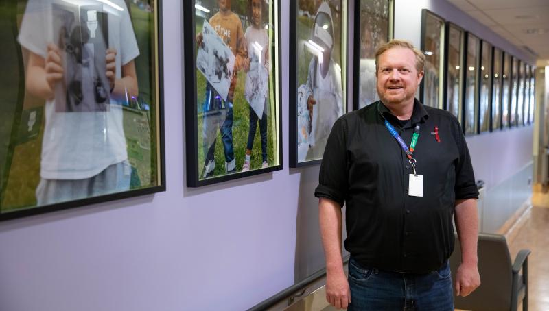 Patient Care Manager, John Balcom, stands and smiles in a light purple hospital hallway beside a series of framed photos on the NICU Hero Wall at Mount Sinai Hospital.