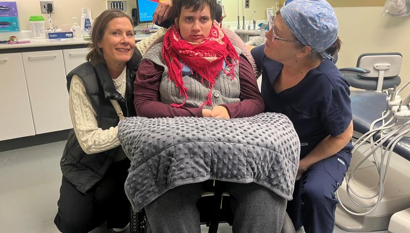 Young woman sits in a wheelchair in a dental clinic, with her dentist and mother smiling beside her