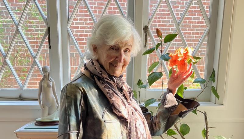 elderly woman stands by a window holding a flower