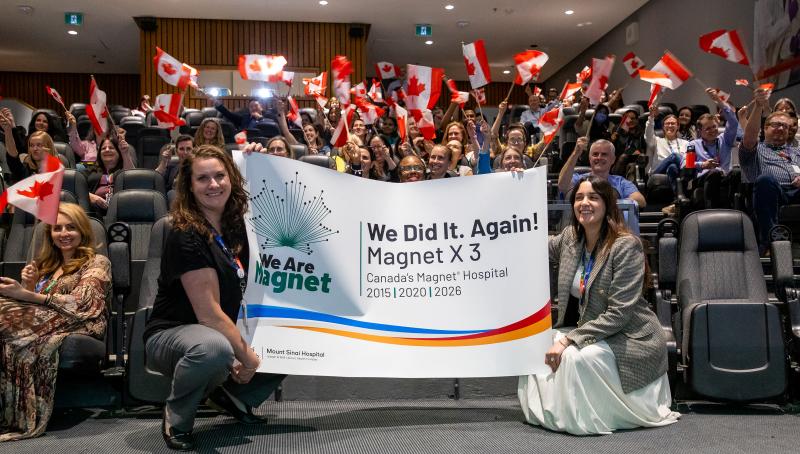 Two people holding a banner celebrating the Magnet recognition in an auditorium with people cheering behind them waving Canadian flags.
