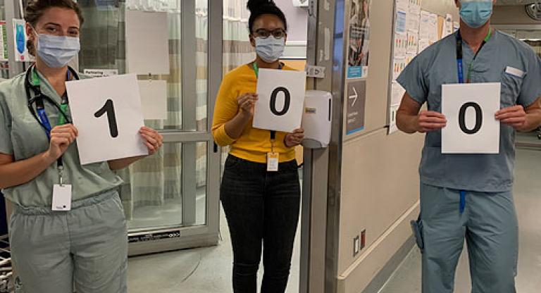 Three hospital staff holding up signs spelling out the number 100"