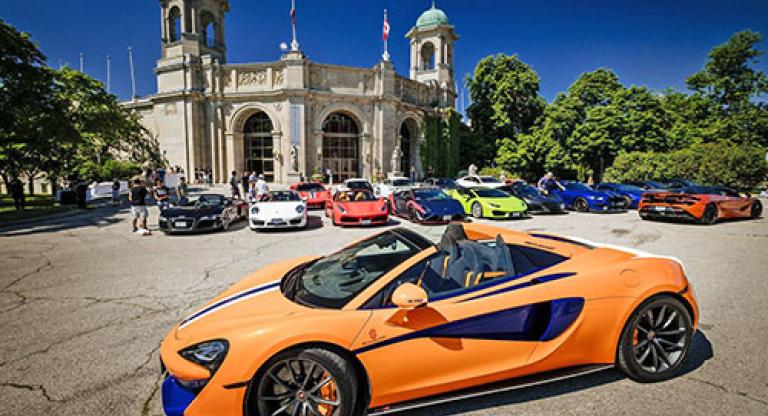 Bright orange luxury car sits in front of other luxury cars and a historic building