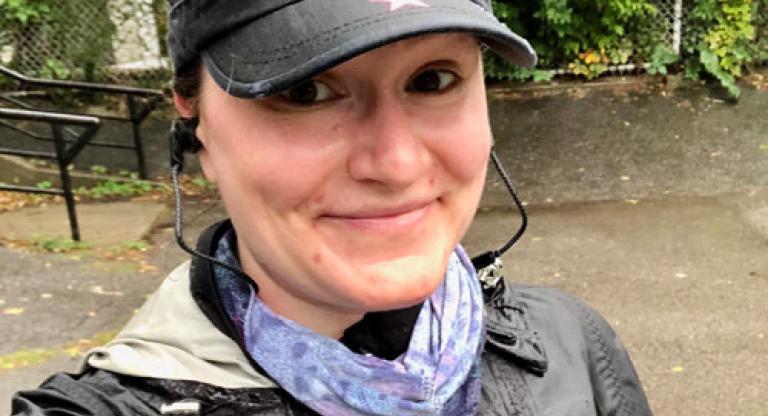Woman stands outside, wearing a hat and smiling at the camera