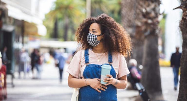 Woman stands outside, holding a coffee cup and wearing a mask