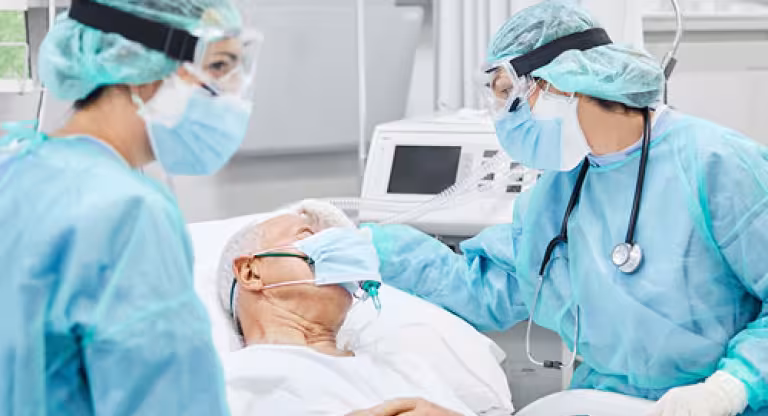 Two health care providers, wearing blue scrubs stand with a patient in the ICU 